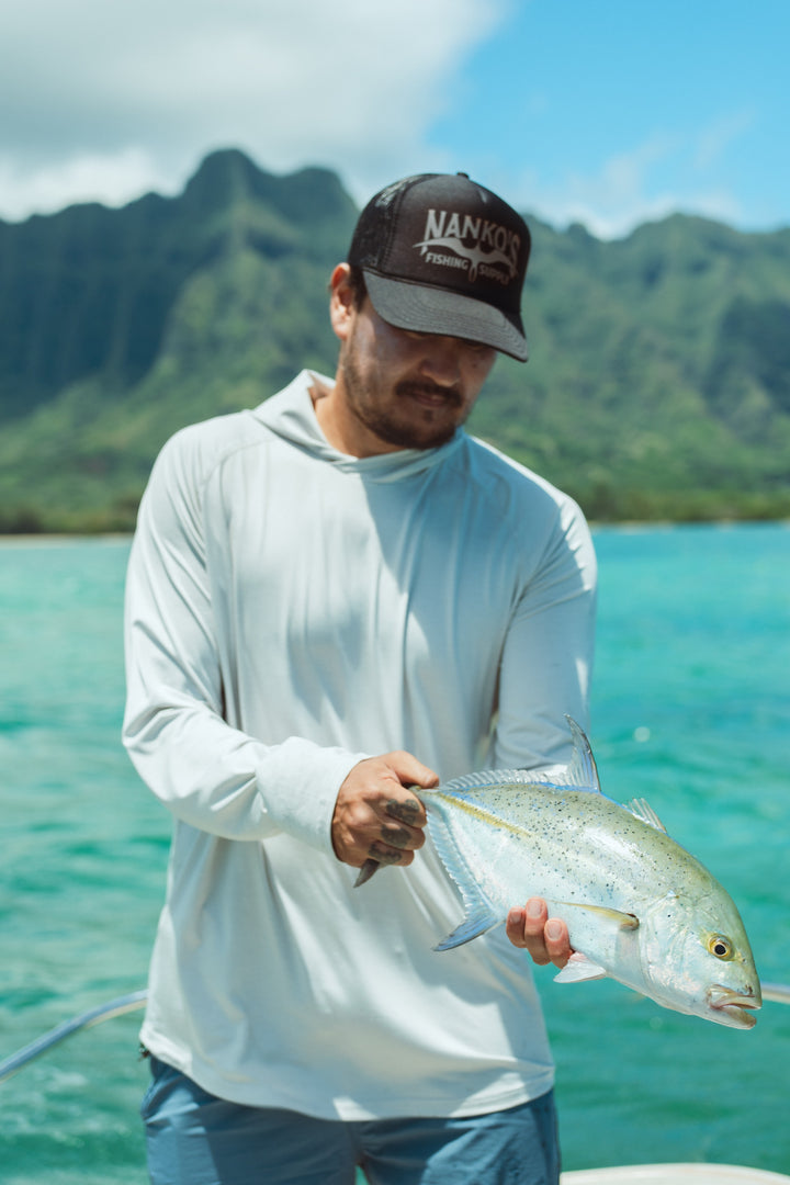Man wearing a Waihana Mariner Storm Hooded Long Sleeve sun shirt holds a fresh-caught fish on a boat in Kaneohe Bay, Hawaii, with turquoise water and dramatic green mountains behind him.